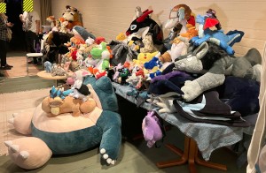 many plushies on a table and on the floor: e.g. IKEA Blåhaj, Pikachu, Snorlax, Angry Bird, Shiron the Windragon (large one with aviator hat)