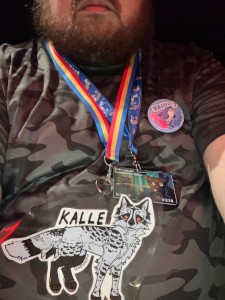 a hairy 40-year-old white man named Kalle with a grey feral canine fursona badge, an IKEA Blåhaj pin, an Animus card, and a grey camo T-shirt