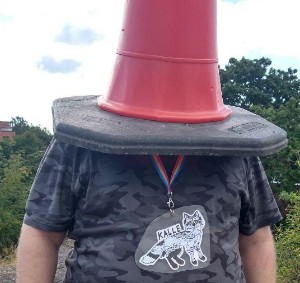 a white 40-year man in a grey camo T-shirt, with a traffic cone on his head, and a drawing of his fursona, a feral black/light grey canine, on his neck